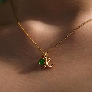 Puede incluir: Un collar de cadena dorada con una piedra preciosa verde y un colgante con la letra 'R' en cursiva.