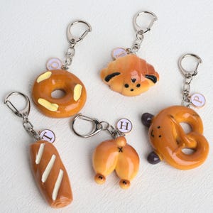 May include: Five food-themed keychains. A glazed donut with yellow icing, a croissant with black accents, a loaf of bread with white stripes, a pair of buns, and a pretzel with purple accents. Each keychain has a silver keyring.