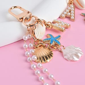 May include: Gold-tone keychain with seashell, pearl, and starfish charms. The charms include a gold scallop shell, a blue starfish, and a white seashell. A strand of pearls hangs from the keychain. The background is pink.