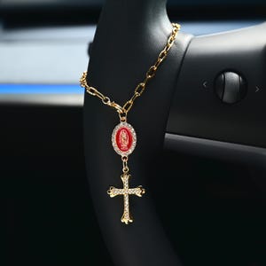May include: Gold-colored car rosary with a red oval pendant featuring a religious image, surrounded by clear crystals. The rosary also includes a gold cross pendant with crystal accents, hanging from a gold chain.