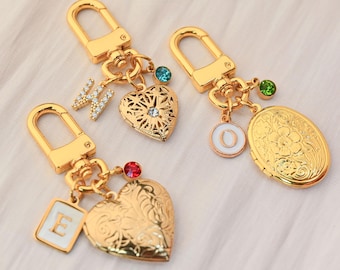 Könnte beinhalten: Goldfarbene Schlüsselanhänger mit verschiedenen Anhängern. Ein Schlüsselanhänger hat ein herzförmiges Medaillon mit floralen Details, ein anderer hat einen herzförmigen Anhänger mit einem Kristall und der dritte hat einen quadratischen Anhänger mit dem Buchstaben "E".