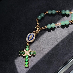 May include: A gold-toned rosary with green beads and a cross pendant. The cross is adorned with green and clear crystals. The pendant features a blue and gold religious emblem. The rosary is displayed on a dark surface.