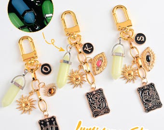 Könnte beinhalten: Goldfarbene Schlüsselanhänger mit verschiedenen Anhängern, darunter ein leuchtender grüner Stein, Sonnen und Tarotkarten-Anhänger. Die Schlüsselanhänger haben auch einen schwarz-goldenen Buchstabenanhänger und einen kleinen, runden Anhänger. Der Text "Luminous Stone" ist im Bild.