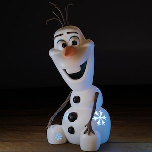 May include: A white Olaf figurine with a carrot nose, black coal buttons, and twig arms. The snowman has a wide smile and a snowflake design on the base. The figurine is a character from the movie Frozen.