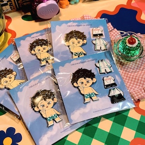 Może przedstawiać: Zestaw pięciu emaliowanych pinów przedstawiających postać z kreskówki w różnych strojach. Postać ma czarne włosy, zielone oczy i jasną karnację. Piny są zapakowane w przezroczyste plastikowe torebki z białym tłem i nadrukiem "Oikawa Dress Up Party".