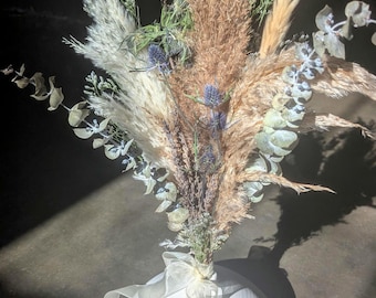 Può includere: Un bouquet di erba della pampa secca, eucalipto e cardi blu disposti in un vaso di ceramica bianca con un nastro di raso bianco.