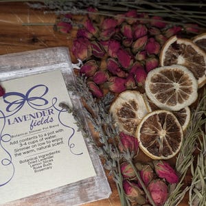 May include: A sheer sachet labeled Lavender Fields Botanical Simmer Pot Blend with instructions to add contents to 3-4 cups of water. The sachet is surrounded by dried lavender sprigs, pink rose buds, and dried lemon slices on a wooden surface.