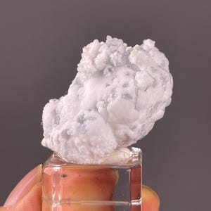 May include: A cluster of white, opaque, and translucent mineral crystals with a bumpy, botryoidal texture. The mineral specimen is mounted on a small, clear, square acrylic base, providing a stable display for the irregular, rounded white formation.