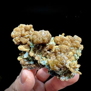Könnte beinhalten: Eine Ansammlung von goldbraunen, botryoidalen Mineralkristallen mit einer durchscheinenden, kristallinen Textur. Das Exemplar weist abgerundete, kugelförmige Formationen auf, die mit kleineren, gezackten Kristallstrukturen und blassgrünen Matrixanteilen durchsetzt sind.