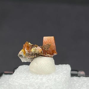 May include: A raw mineral specimen featuring translucent, amber-coloured wulfenite crystals with sharp, tabular edges. The cluster displays a mix of deep orange and golden hues, attached to a small, textured, light-coloured matrix base.