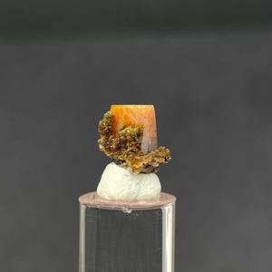May include: A small, orange, prismatic mineral crystal specimen featuring a cluster of dark olive-green, granular inclusions attached to one side. The crystal is mounted on a white, textured adhesive base atop a clear, cylindrical acrylic display stand.