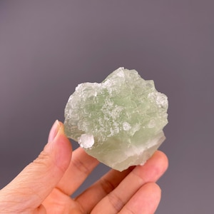 Puede incluir: Un racimo de cristales de fluorita de color verde pálido con una estructura translúcida, geométrica y cúbica. El espécimen mineral muestra un tono menta claro con bordes claros y afilados y una textura superficial cristalina que capta la luz.
