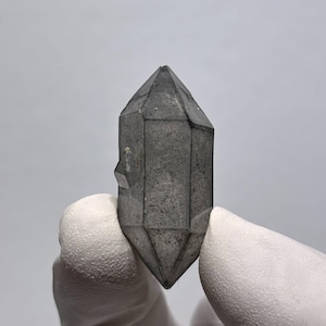 May include: A double-terminated smoky quartz crystal with a translucent, dark gray, and charcoal-colored appearance. The mineral specimen features distinct geometric facets and a sharp, pointed termination at both the top and bottom ends.