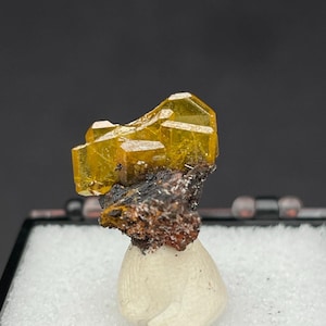 May include: A cluster of translucent, golden-yellow wulfenite crystals sits atop a dark, textured matrix of iron-rich mineral. The specimen is mounted on a small, smooth, off-white, rounded base, providing a stable display for the mineral.