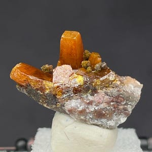May include: A wulfenite mineral specimen featuring vibrant orange, tabular crystals protruding from a textured, multi-coloured matrix base. The crystalline structure displays sharp edges and a glossy finish, with small clusters of green mineral deposits.