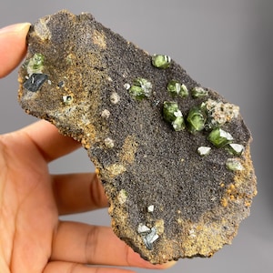 May include: A dark, textured rock formation with several vibrant green crystals and smaller white and dark crystals. The rock has a rough, porous surface with areas of brown and gold coloration. The crystals are clustered across the surface.