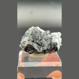May include: A raw mineral specimen featuring dark, metallic, crystalline structures with sharp, angular facets. The cluster displays a high-luster, charcoal-gray surface contrasted by a patch of white, granular mineral deposits on one side.