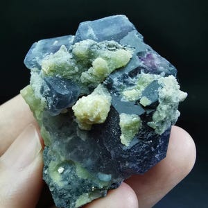 Può includere: Un esemplare minerale grezzo caratterizzato da cristalli di fluorite cubici, traslucidi e di colore viola intenso, raggruppati con depositi minerali testurizzati di colore giallo-verde pallido. La struttura cristallina presenta bordi geometrici netti.