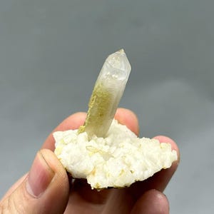 May include: A clear, pointed quartz crystal cluster featuring a single prominent vertical point with a pale yellow-green mineral inclusion at its base, set upon a textured, opaque white crystalline matrix with jagged, irregular edges.