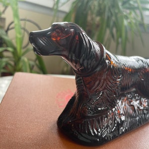 May include: A dark brown glass figurine of a dog, possibly a hunting dog, with a detailed design. The dog is standing on a base with a textured surface.