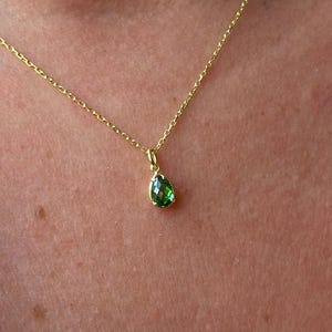 Könnte beinhalten: Eine zarte goldene Halskette mit einem kleinen, birnenförmigen Anhänger aus grünem Edelstein. Der Anhänger hängt an einer feinen Goldkette. Die Halskette wird vor einem Hautton-Hintergrund gezeigt.