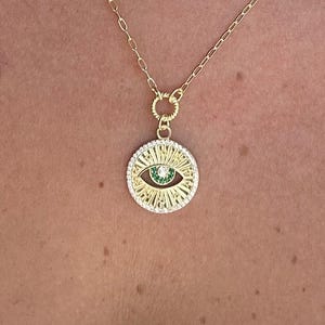 Könnte beinhalten: Goldfarbene Halskette mit einem kreisförmigen Anhänger mit einem Augendesign. Der Anhänger hat eine grüne Mitte, umgeben von weißen Steinen und einem Sonnenstrahlenmuster. Die Halskette wird von einer Person getragen.