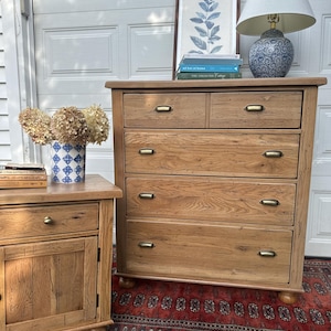 May include: A light-colored wooden dresser with five drawers and brass hardware. A matching nightstand is to the left. A blue and white patterned lamp sits on top of the dresser, along with a framed botanical print and books. A red rug is beneath the dresser.