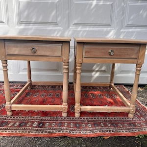 May include: Two matching wooden side tables with a single drawer and turned legs. The tables are light brown and sit on a red patterned rug. Each drawer has a round brass knob.
