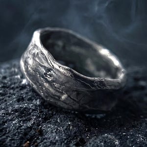 May include: A silver ring with a highly textured, uneven surface and a dark, oxidized finish. The band features organic, rugged contours that mimic natural rock formations, creating a distinct, weathered appearance throughout the metal.