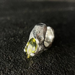 May include: A silver ring with a textured, rugged surface featuring a single marquise-cut lime green gemstone set into the metal. The band has an irregular, organic shape with a metallic finish that contrasts with the faceted, bright green stone.