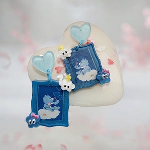 May include: Blue Care Bear earrings with a heart-shaped stud. Each earring features a blue frame with a Care Bear illustration on a cloud. Additional embellishments include blue hearts, white clouds, and blue star-shaped charms.