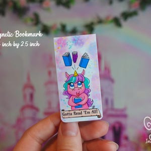 May include: A pink unicorn with a blue and purple mane and tail is holding a book and surrounded by books. The text on the bookmark reads "Gotta Read 'Em All!"