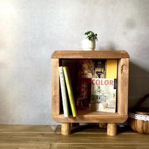 Puede incluir: Una estantería de madera con un estante abierto cuadrado y cuatro patas. La estantería contiene tres libros, uno de los cuales se titula "Interiors in Color". Una pequeña taza blanca con una planta verde está encima de la estantería.