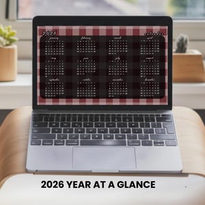 Pode incluir: Um laptop exibe um calendário de 2026 com um padrão xadrez bordô e branco. O calendário mostra todos os doze meses, com o texto "2026 YEAR AT A GLANCE" na parte inferior. O laptop está em uma superfície de madeira clara.