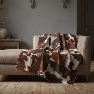 Könnte beinhalten: Eine braun-weiße Patchwork-Decke aus Kuhfell, die über einem beigen Sofa drapiert ist. Die Decke weist eine Mischung aus braunen und weißen Flecken auf, die ein rustikales und strukturiertes Aussehen erzeugen. Der Überwurf verleiht dem Wohnzimmer einen Hauch von Wärme und Stil.