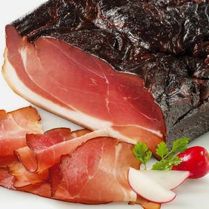 May include: A close-up of thinly sliced smoked ham, with a larger piece of ham in the background. The ham is a deep red color and has a slightly shiny surface. The ham is arranged on a white surface with a few sprigs of parsley and a small red radish.