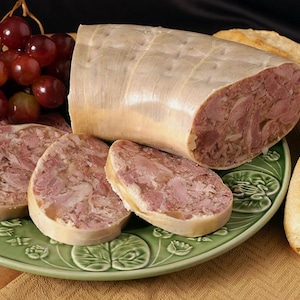 May include: A close-up of a platter of sliced head cheese, a type of meatloaf made from pork head and other meats. The slices are arranged on a green and white patterned plate, with a whole log of head cheese in the background. There are also some grapes in the background.