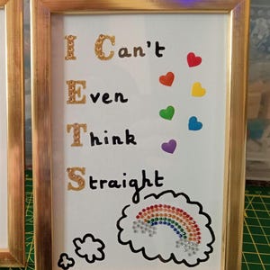 May include: A gold-framed wall art piece featuring the text I Can't Even Think Straight in black lettering, with the letters I, E, T, and S accented in gold glitter. A colorful rainbow made of small rhinestones sits inside a black cloud outline.