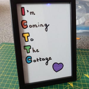May include: A black rectangular frame containing a white sign with the hand-lettered text I'm Coming To The Cottage. The first letter of each word is highlighted in a different color: red, orange, yellow, green, and blue. A purple heart is drawn below.