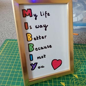 May include: A gold-colored rectangular picture frame containing a white card with the handwritten text: My life Is way Better Because I met You, with the first letter of each line in a different color and a red heart symbol at the bottom.