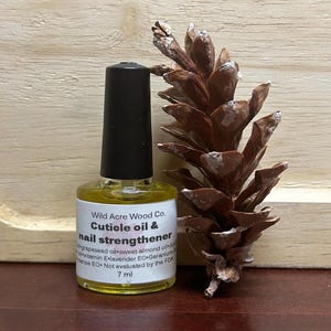 May include: A small, clear glass bottle with a black cap, containing a yellow liquid. The bottle is labelled "Wild Acre Wood Co. Cuticle oil & nail strengthener" and lists the ingredients as grapeseed oil, sweet almond oil, vitamin E, lavender EO, and geranium EO. The bottle is sitting on a wooden surface next to a brown pine cone.
