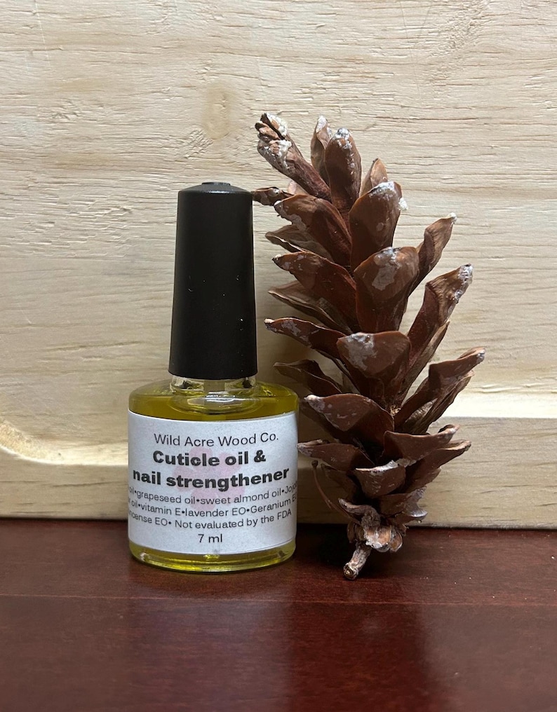 May include: A small, clear glass bottle with a black cap, containing a yellow liquid. The bottle is labelled "Wild Acre Wood Co. Cuticle oil & nail strengthener" and lists the ingredients as grapeseed oil, sweet almond oil, vitamin E, lavender EO, and geranium EO. The bottle is sitting on a wooden surface next to a brown pine cone.