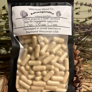 May include: A black resealable bag with a white label that reads "Wild Acre Wood Co. Ashwagandha Withania somnifera USDA organic | GMP certified Size 00 vegetable capsules 50 100 200 Packaged in small batches in Raymond Wisconsin USA". The bag contains a large quantity of beige capsules.
