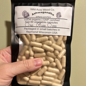 May include: A black resealable bag with a white label that reads "Wild Acre Wood Co. Ashwagandha Withania somnifera USDA organic | GMP certified Size 00 vegetable capsules 50 100 200 Packaged in small batches in Raymond Wisconsin USA". The bag contains a large quantity of light brown capsules.