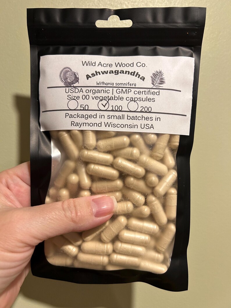 May include: A black resealable bag with a white label that reads "Wild Acre Wood Co. Ashwagandha Withania somnifera USDA organic | GMP certified Size 00 vegetable capsules 50 100 200 Packaged in small batches in Raymond Wisconsin USA". The bag contains a large quantity of light brown capsules.