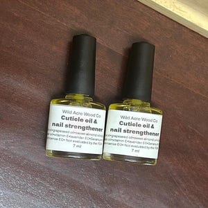 May include: Two 7 ml bottles of cuticle oil and nail strengthener by Wild Acre Wood Co. The bottles are clear and contain a yellow liquid. The labels are white with black text and list the ingredients: grapeseed oil, sweet almond oil, vitamin E, lavender EO, geranium, and incense EO. The bottles are not evaluated by the FDA.