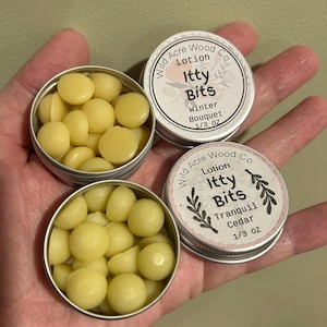 May include: Two small silver tins containing lotion bars. The tins are labeled "Wild Acre Wood Co Lotion Itty Bits" with different scents. The top tin is labeled "Winter Bouquet" and the bottom tin is labeled "Tranquil Cedar". Both tins contain 1/3 ounce of lotion bars.