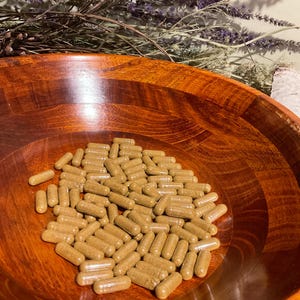 May include: A collection of numerous light brown, cylindrical capsules filled with a granular substance, resting inside a polished, dark reddish-brown wooden bowl with a visible grain pattern and a smooth, rounded interior surface.