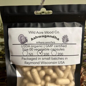 May include: A clear plastic bag with a black border contains a large quantity of beige capsules. The bag has a label that reads "Wild Acre Wood Co. Ashwagandha Withania somnifera USDA organic | GMP certified Size 00 vegetable capsules 50 100 200 Packaged in small batches in Raymond Wisconsin USA".