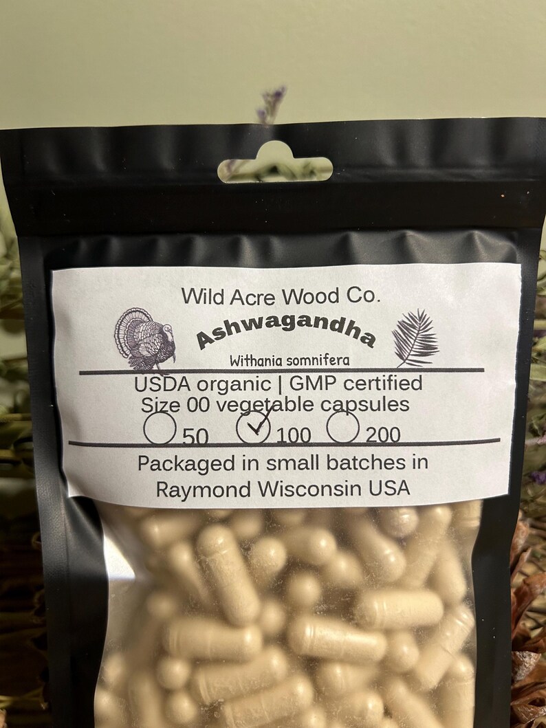 May include: A clear plastic bag with a black border contains a large quantity of beige capsules. The bag has a label that reads "Wild Acre Wood Co. Ashwagandha Withania somnifera USDA organic | GMP certified Size 00 vegetable capsules 50 100 200 Packaged in small batches in Raymond Wisconsin USA".